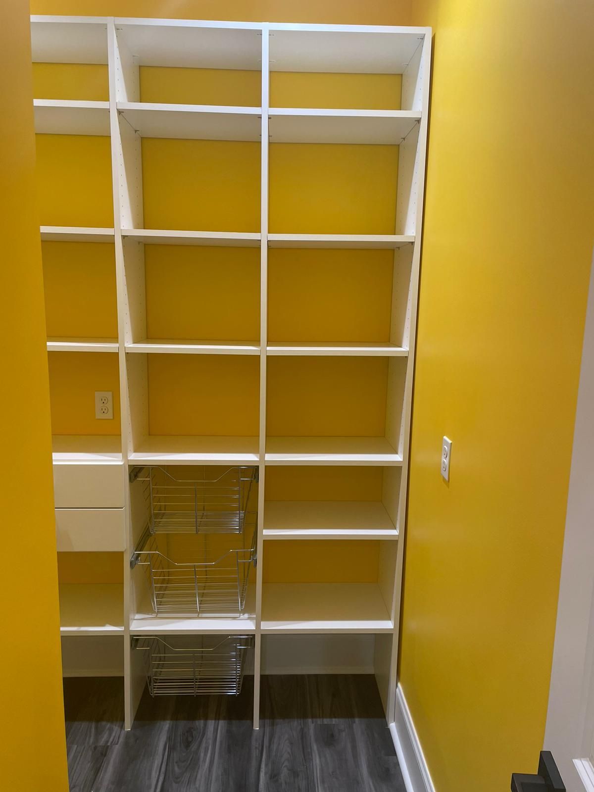 White shelving unit against a bright yellow wall, with pull-out wire baskets.