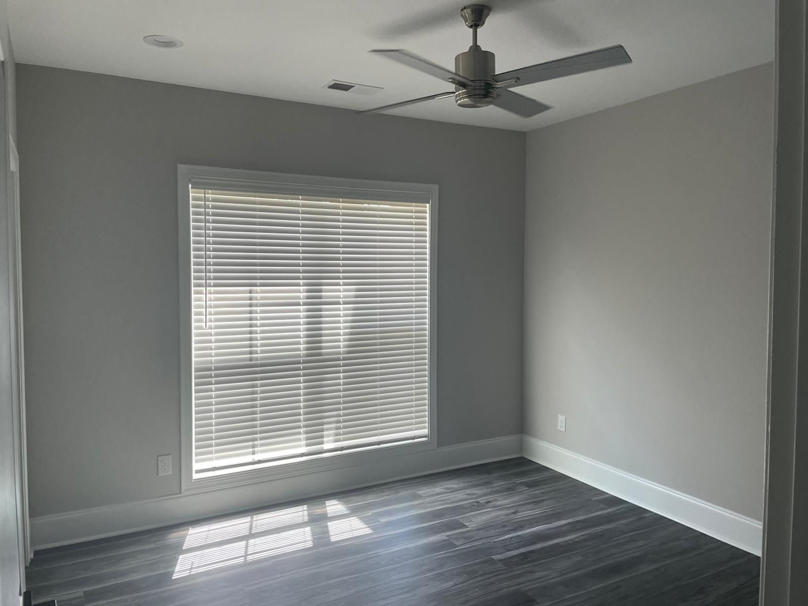 Empty room with a window, blinds, and ceiling fan; dark floor, light gray walls.