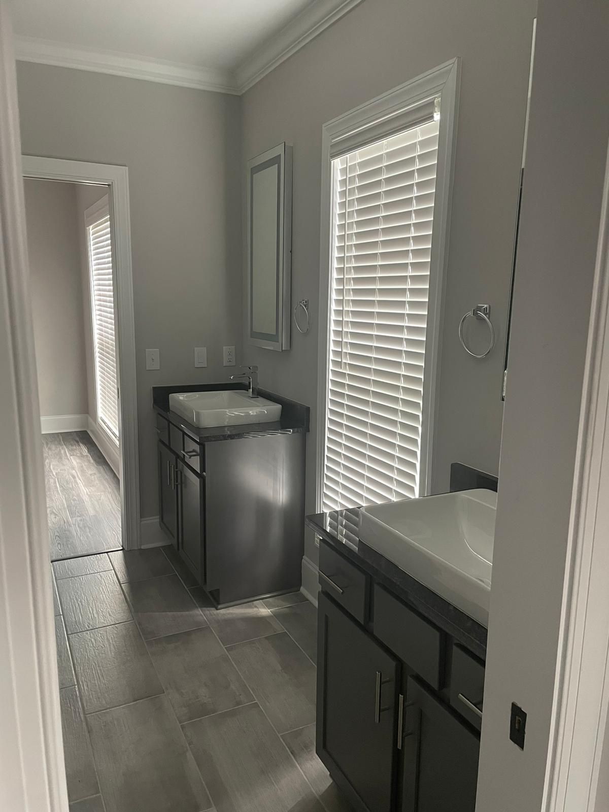 Bathroom with two sinks and gray cabinets; window with blinds; gray tile floor.