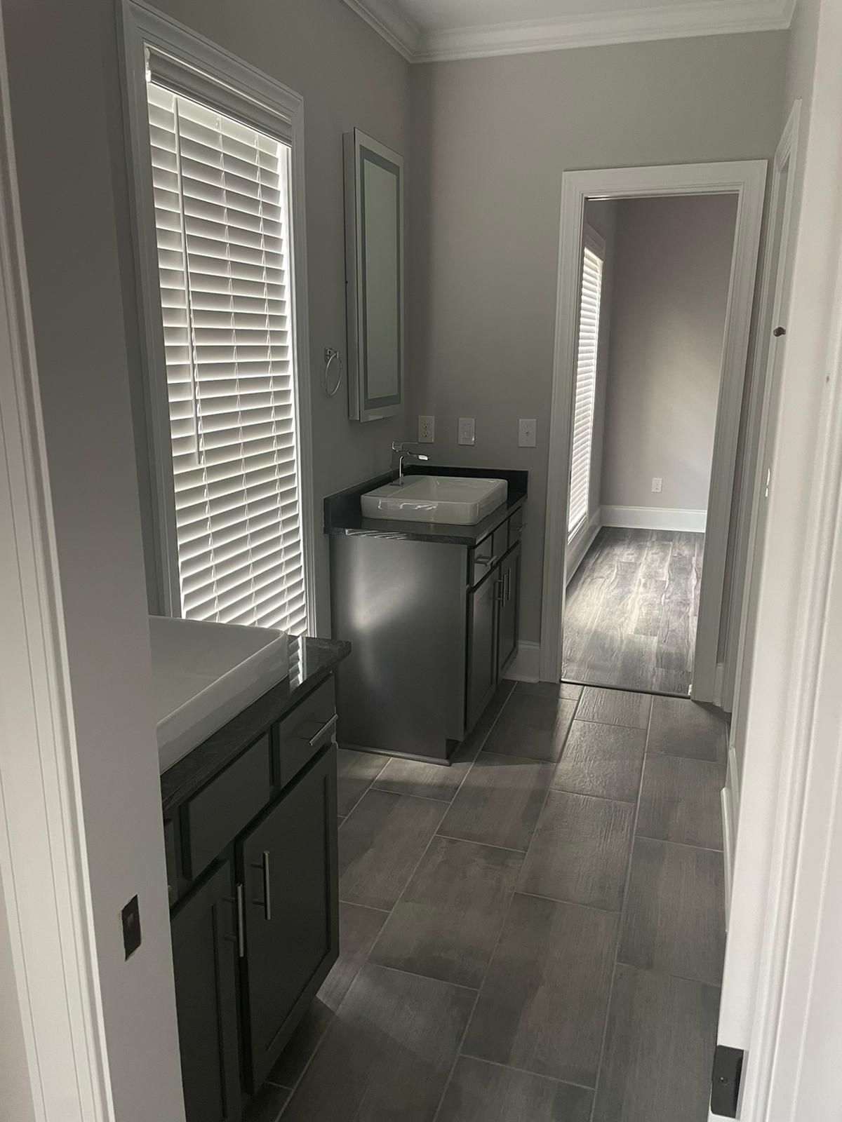 Bathroom with two dark cabinets, sinks, and a doorway to a room with a window. Gray tile floor.