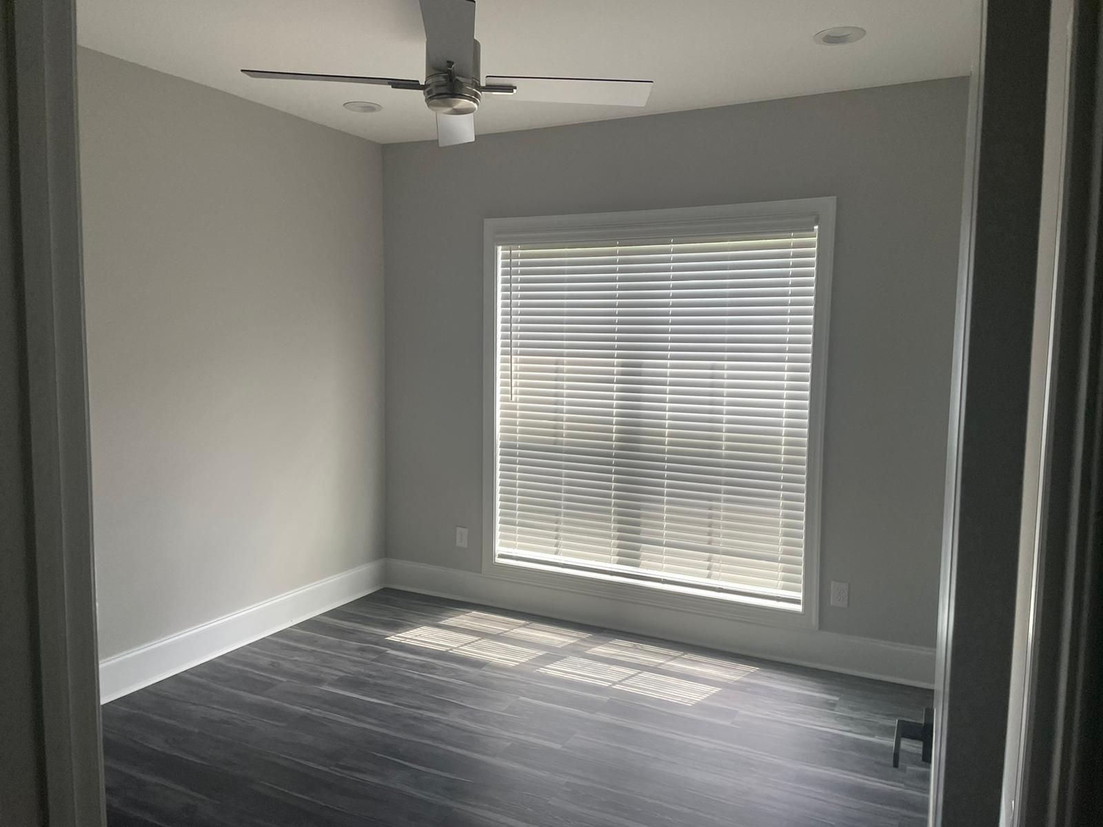 Empty room with gray walls, a window with blinds, dark floor, and a ceiling fan.