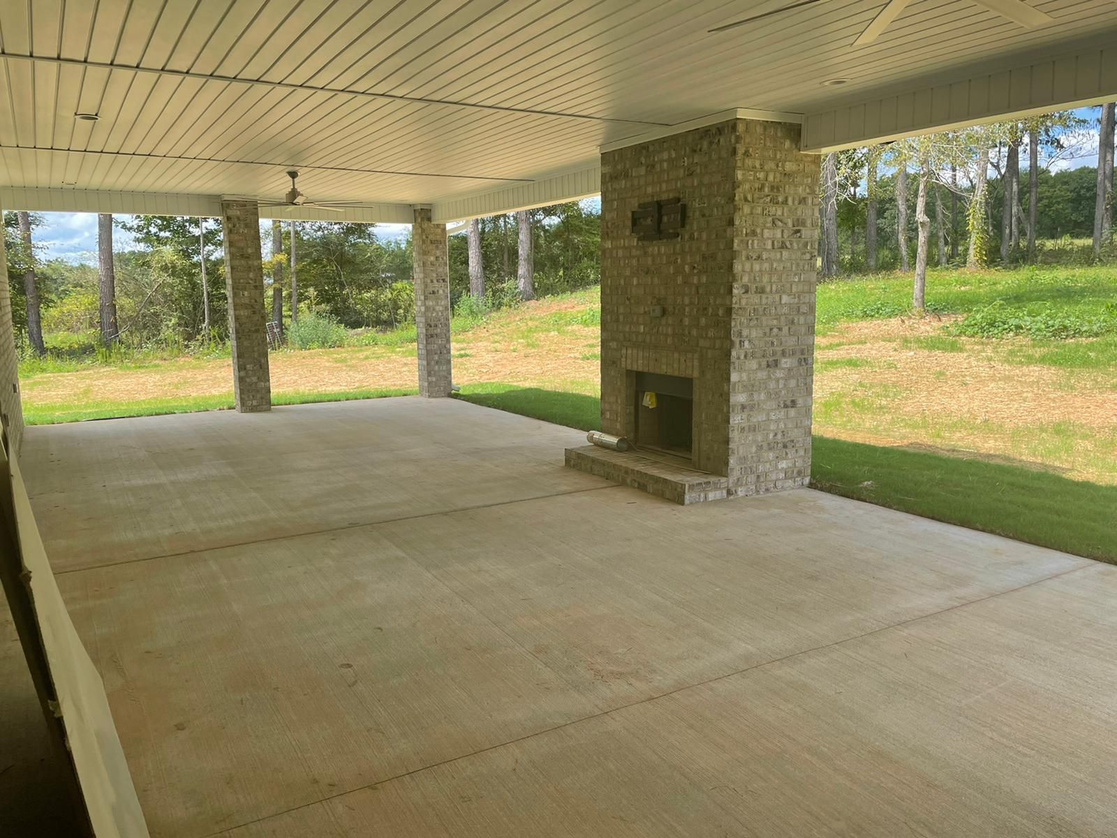 Covered patio with concrete floor, stone fireplace, and pillars overlooking a grassy field.