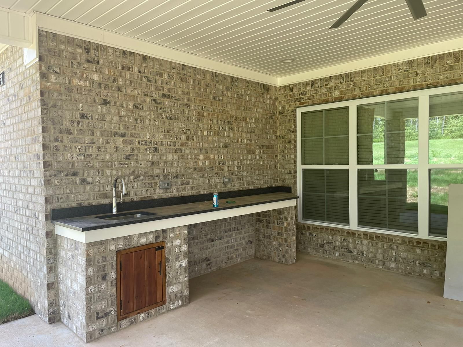 Outdoor kitchen with brick walls, a sink, and a window overlooking greenery.