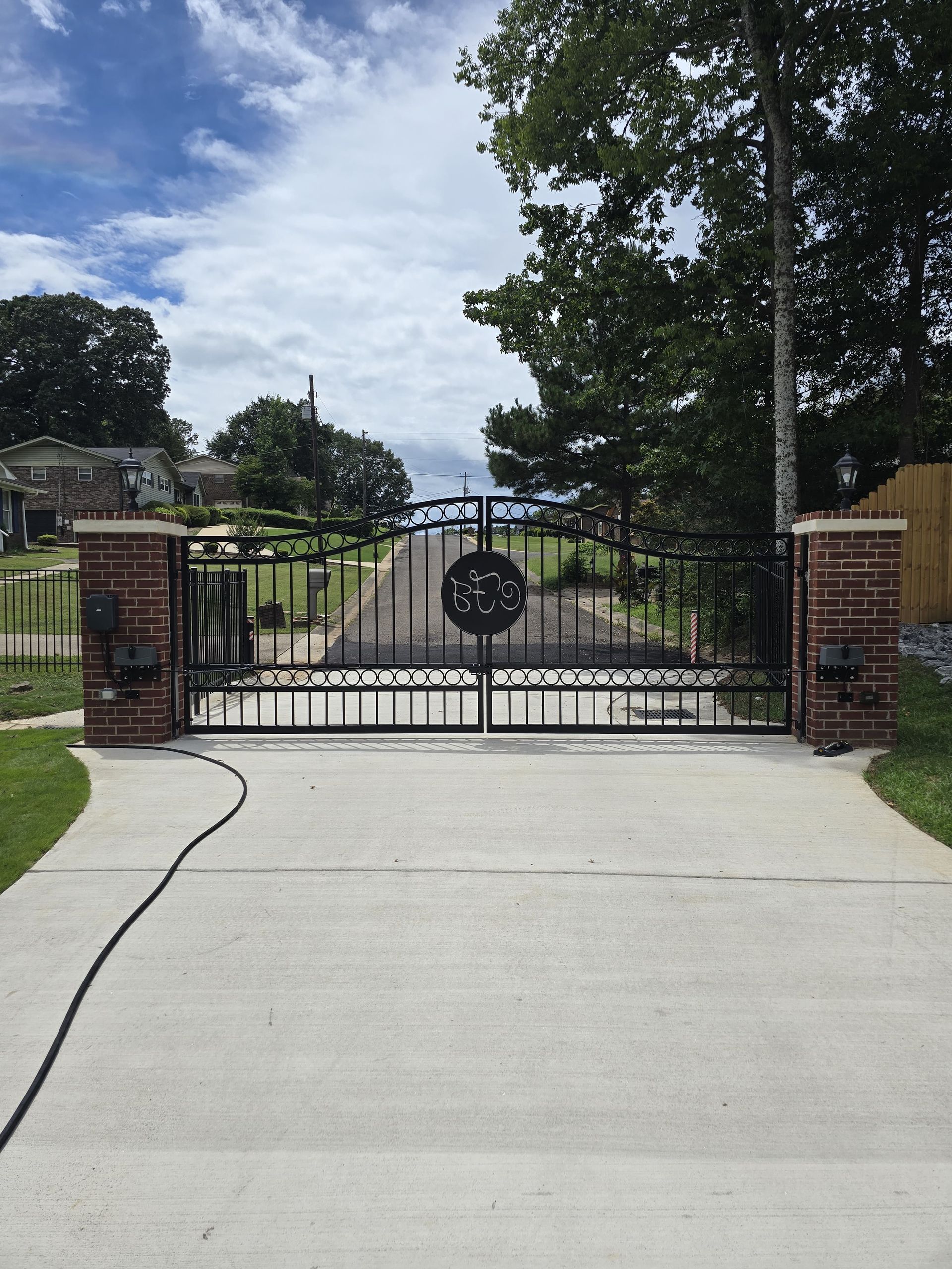 Black metal driveway gate with a circular emblem, set between brick pillars. Concrete driveway leads towards trees and sky.