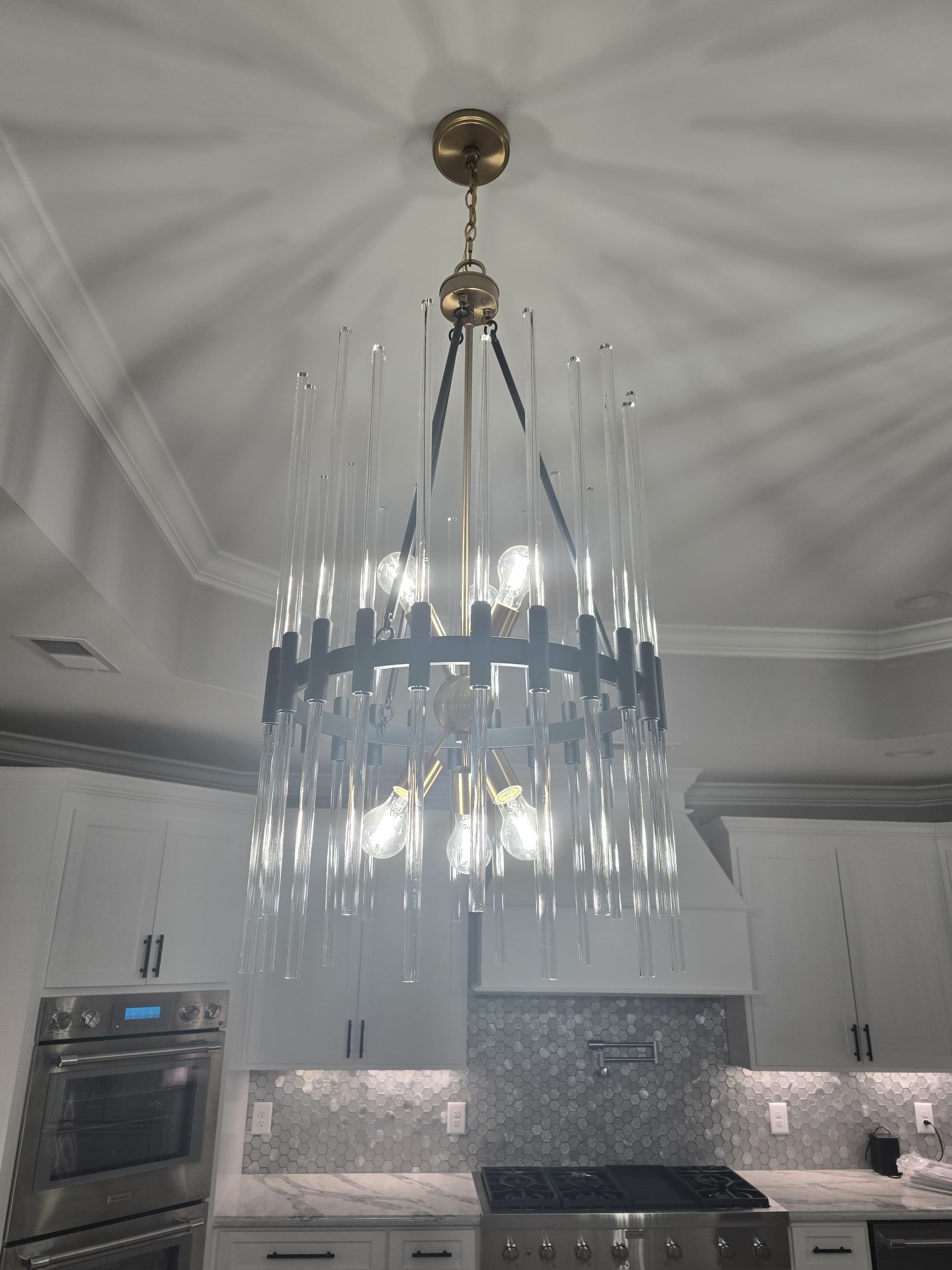 A modern chandelier hangs in a kitchen, illuminating glass rods with gold and black accents.