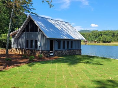 Gray lakeside cabin with a metal roof on a grassy lawn under a blue sky.