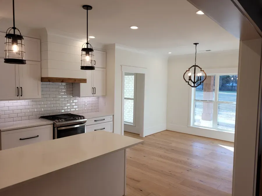 White kitchen with light wood floors, two hanging lights, and a view of a windowed dining area.