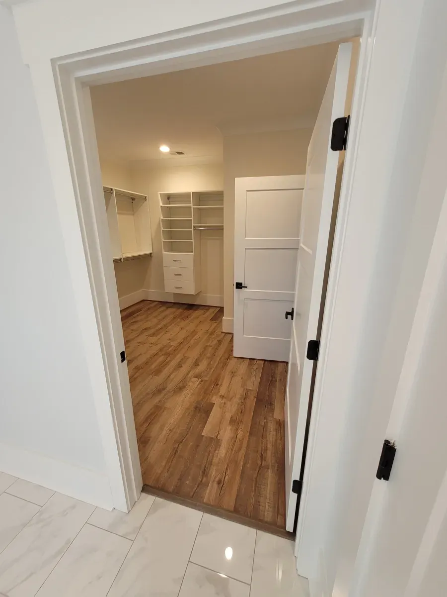 Open doorway to a bright closet with white shelves, a closed white door, and wood flooring.