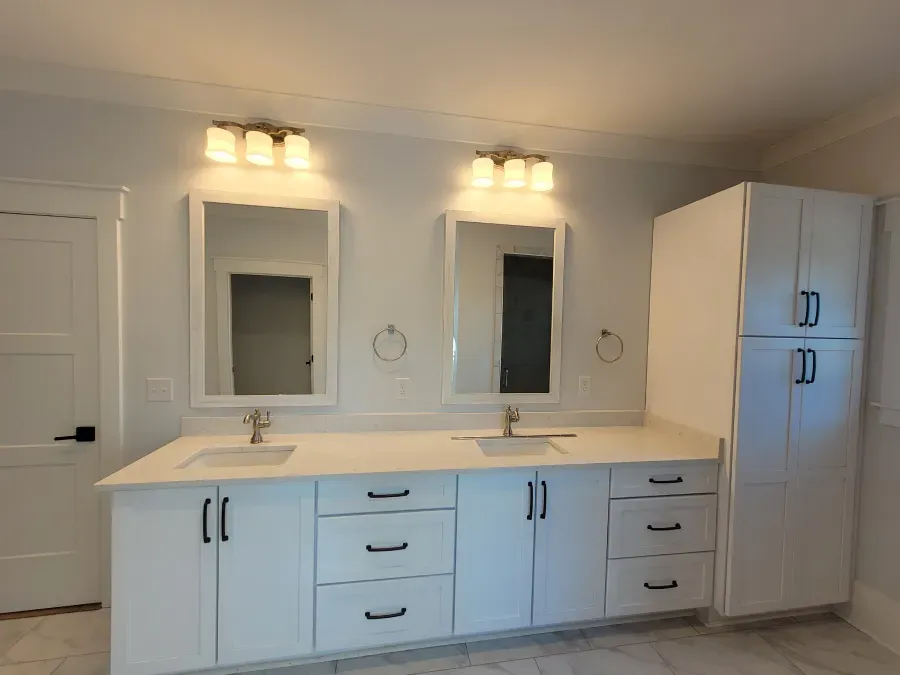 White bathroom with double sinks, mirrors, and a tall cabinet; gold fixtures, black hardware.