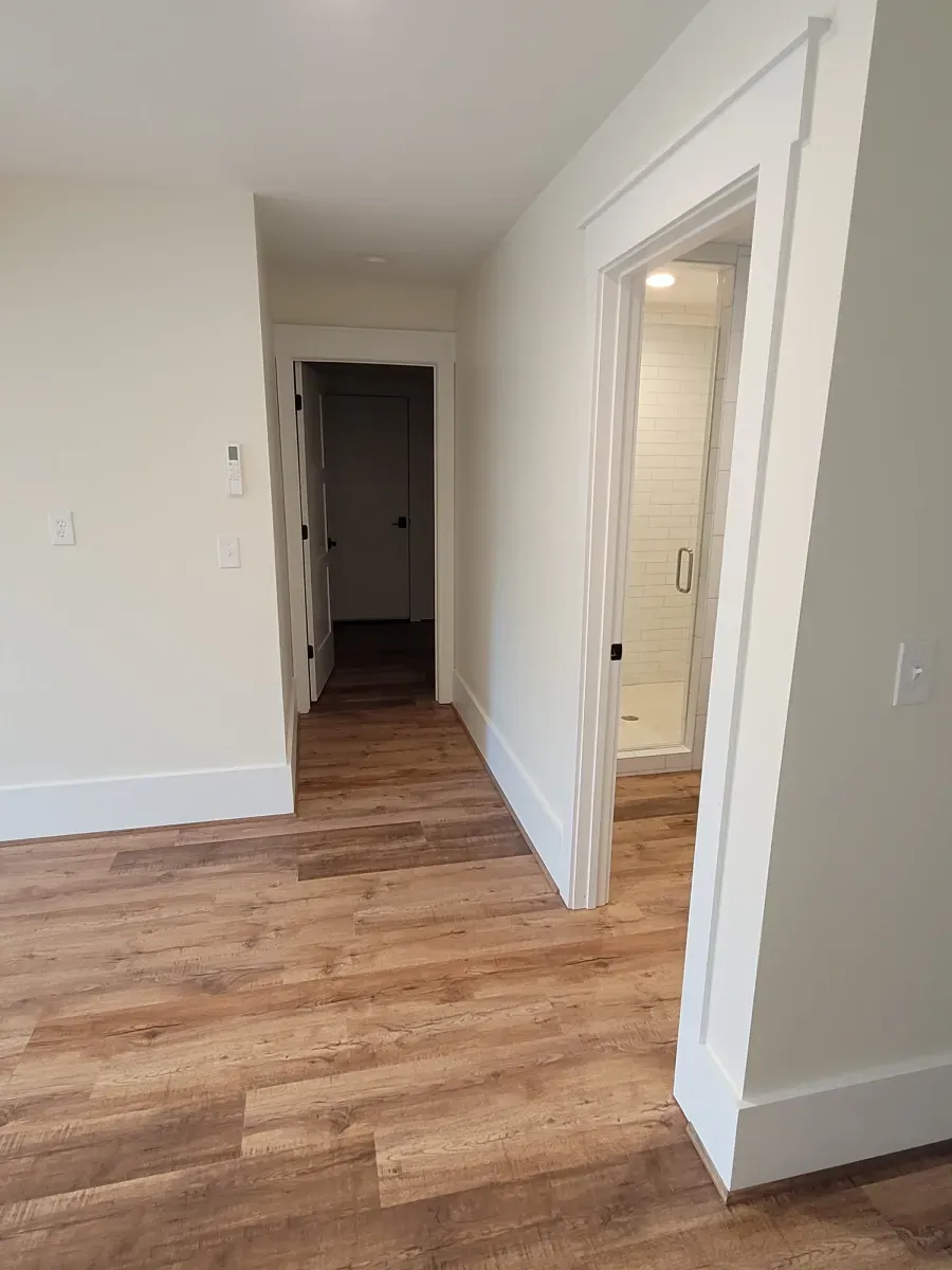 Hallway with wood-look floor, white walls, and open doors to a bathroom and another room.