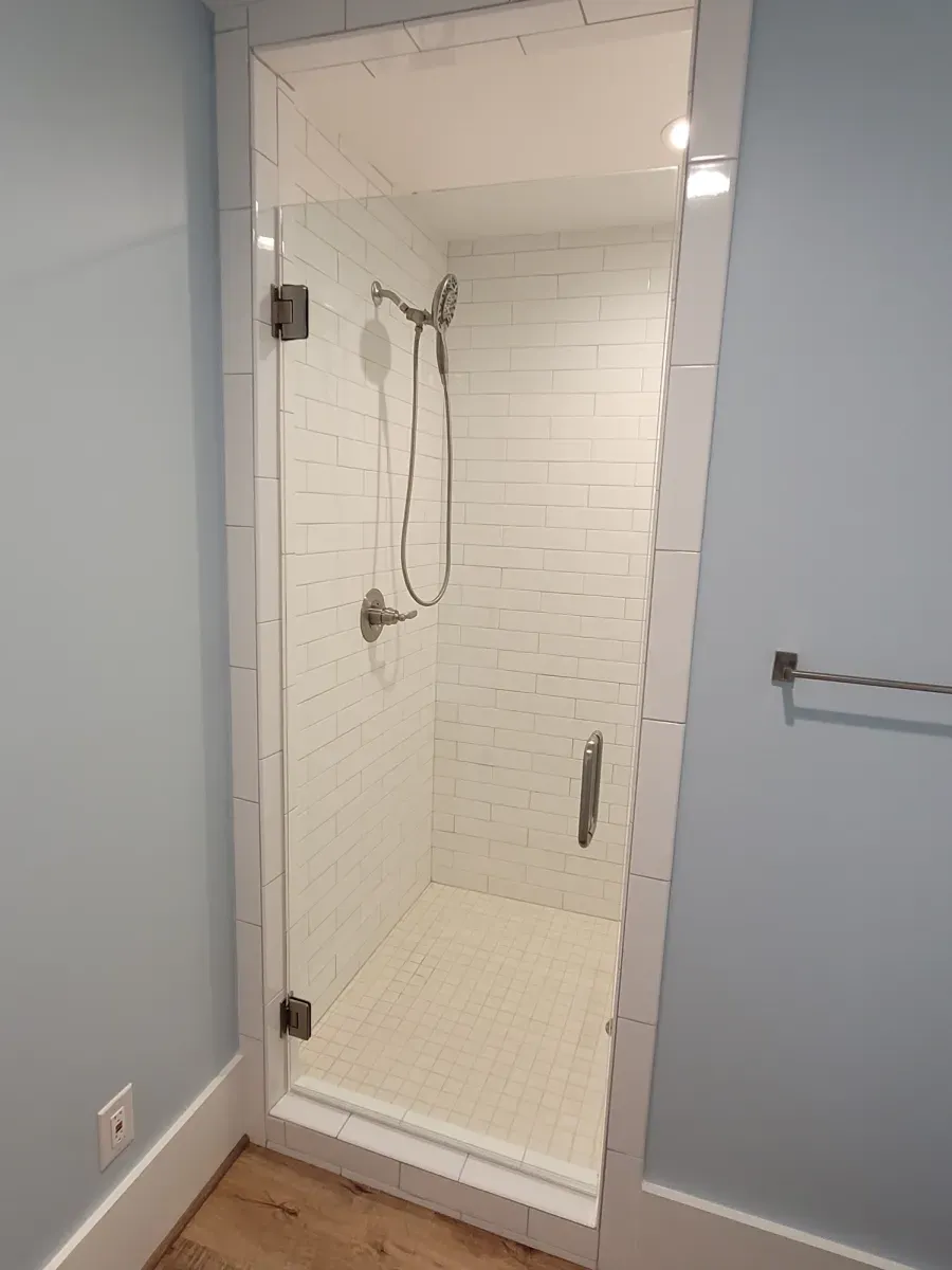Glass shower with white tiled walls and a gray shower head in a blue bathroom.