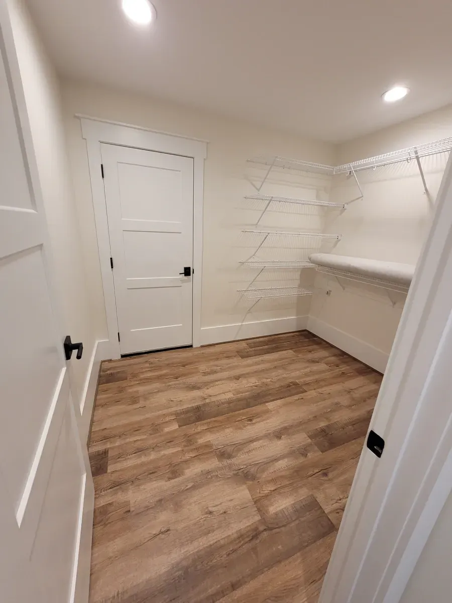 Empty closet with wood-look floor, white shelves, door, and trim, lit by overhead lights.