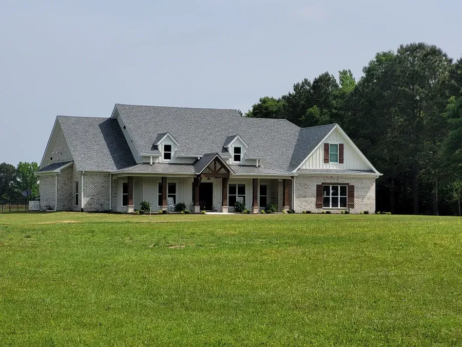 Large, light-colored brick house with gray roof and green lawn in front, trees in the background.