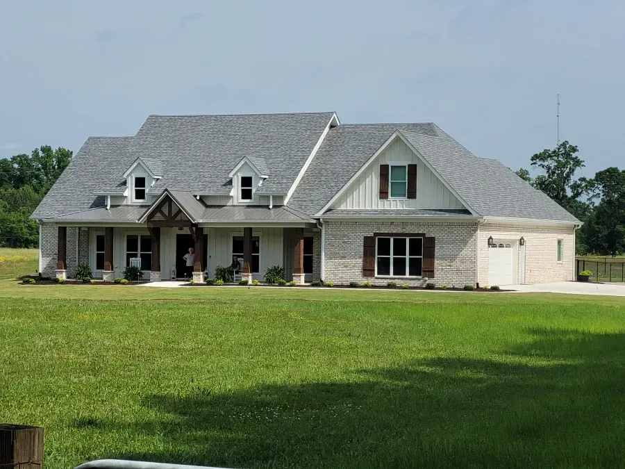 A large, light-colored house with a gray roof, brown shutters, and a green lawn under a blue sky.