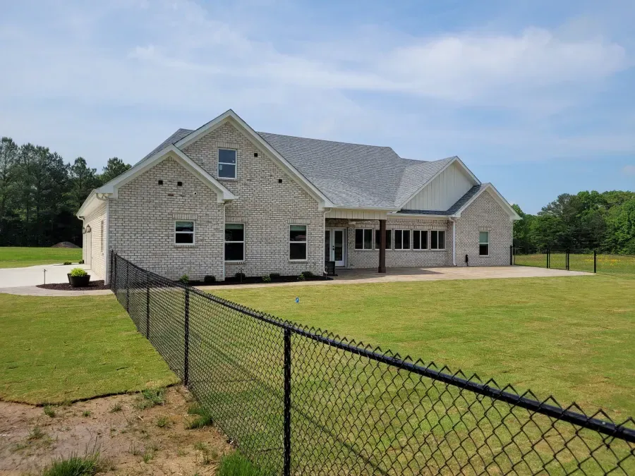 Light brick house with gray roof, black chain-link fence, and green lawn under a blue sky.
