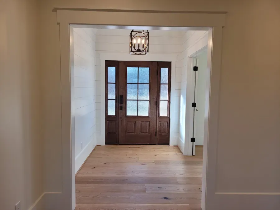 Wooden front door with glass panels inside a white-walled entry with hardwood flooring and a hanging light fixture.