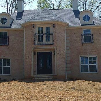 Beige brick house with black roof, door, and railings. Oval windows, chimneys.