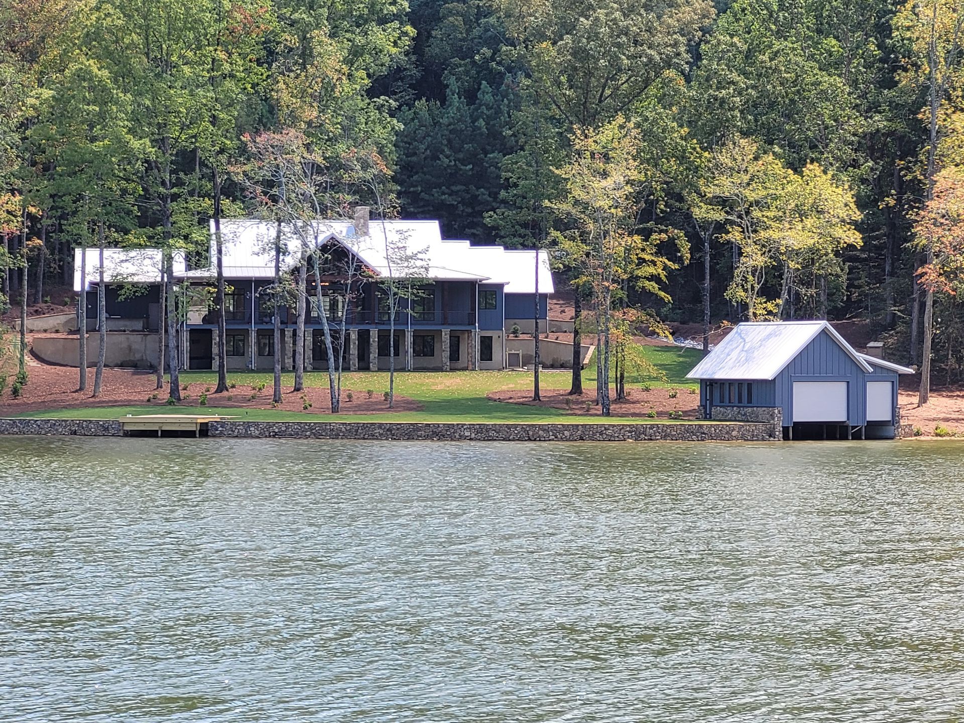 Lakeside home with a light-colored roof, trees, and a boat house on a lake.