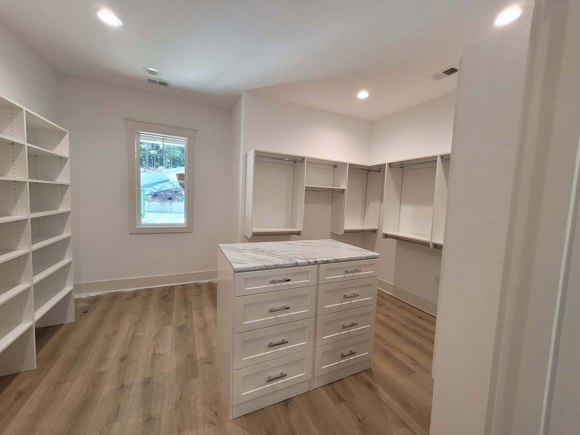 Empty white walk-in closet with island drawers, shelves, and window. Light wood floor.