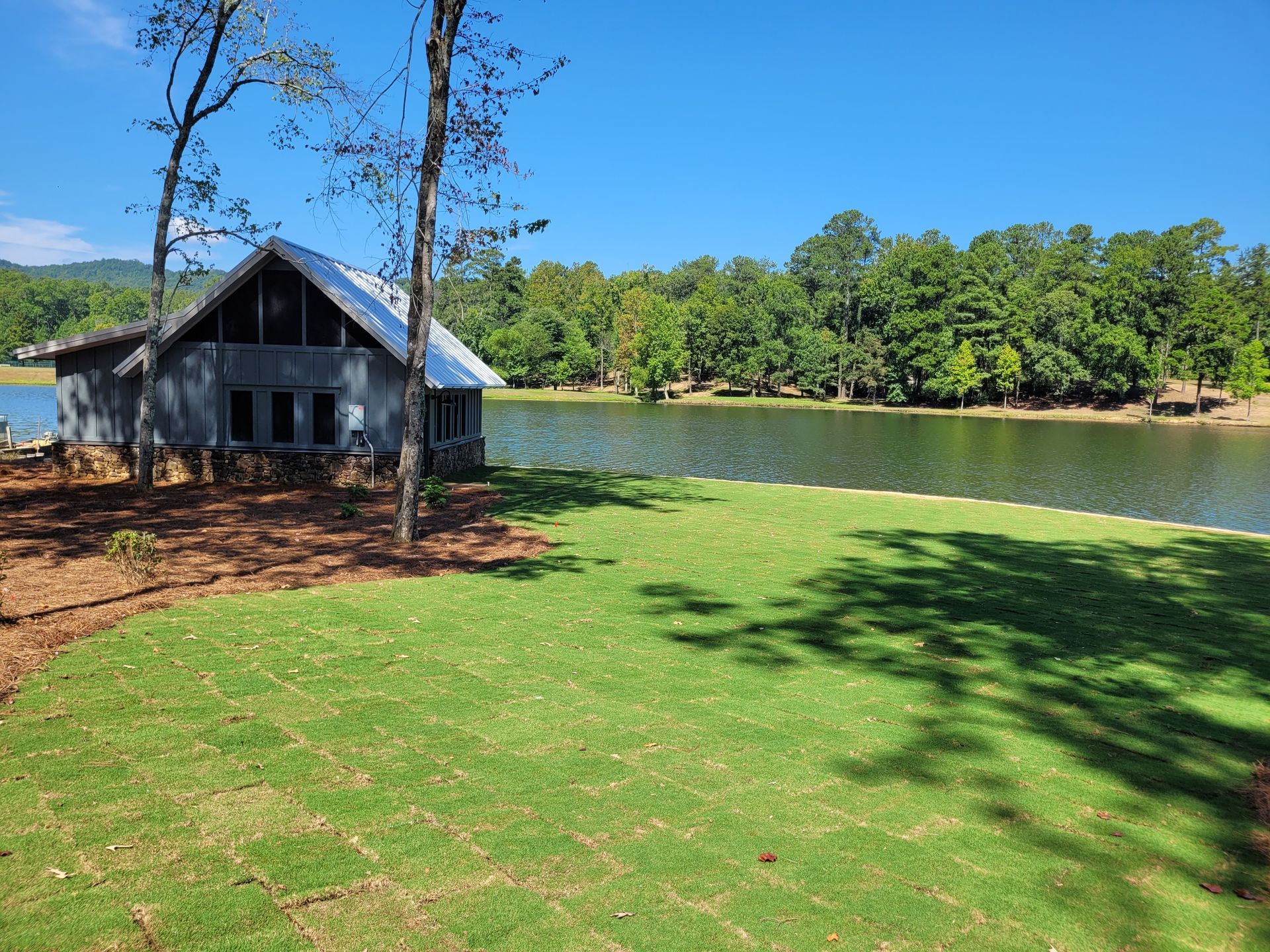 A lakefront cabin with a green lawn, trees, and a bright blue sky.