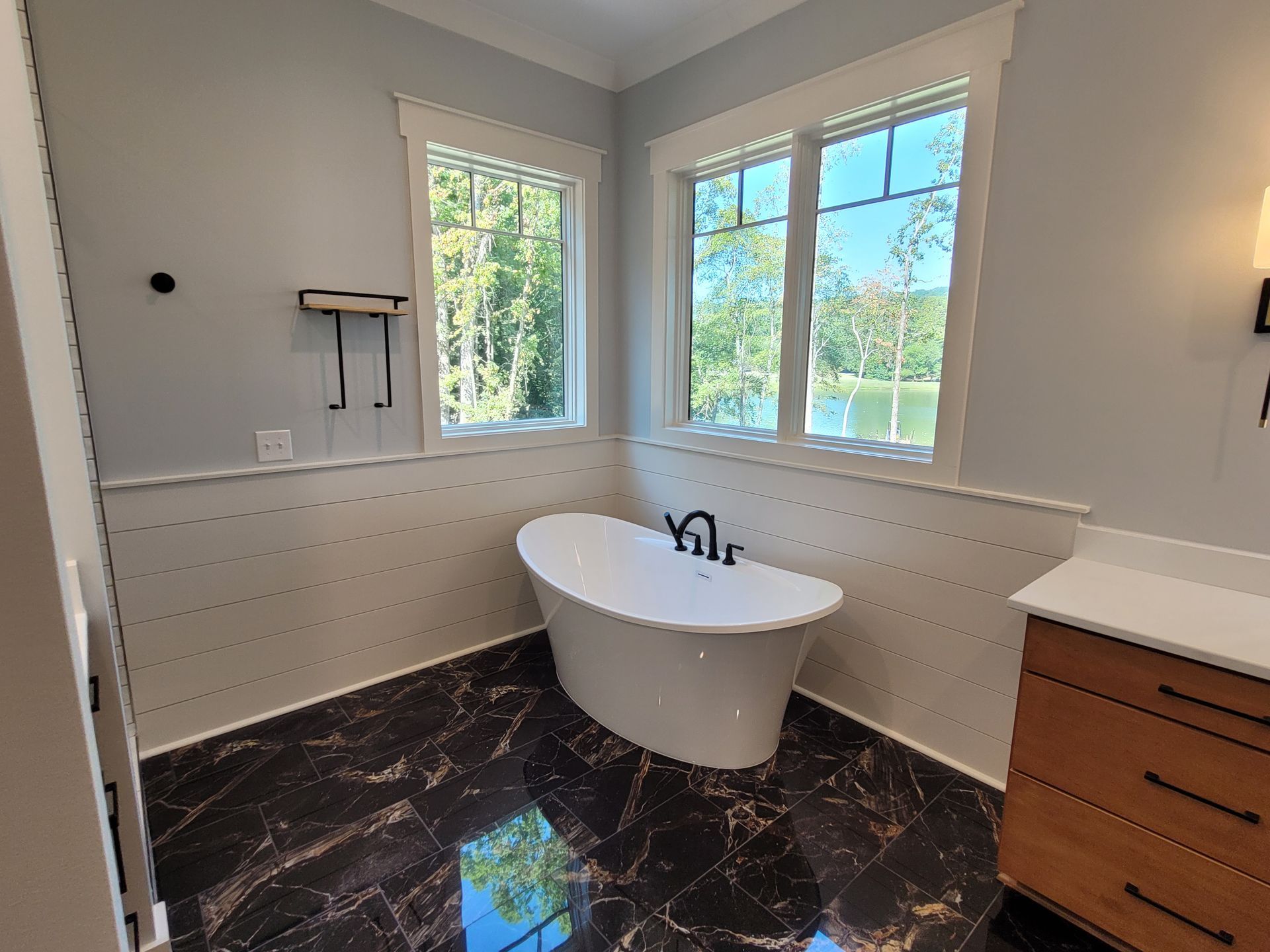 Bathroom with a white oval bathtub by windows overlooking a lake, dark tile floor, and light blue walls.