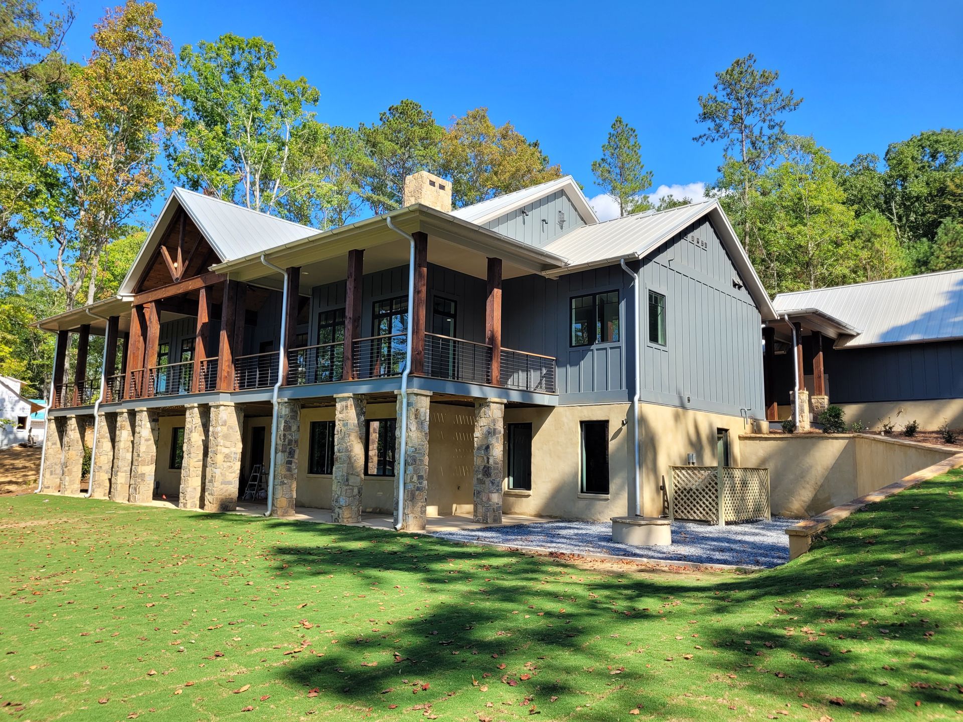 Two-story house with a covered porch, stone pillars, and metal roof, set on a green lawn with trees in the background.