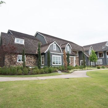 Large gray and stone house with a long driveway on a grassy lawn.