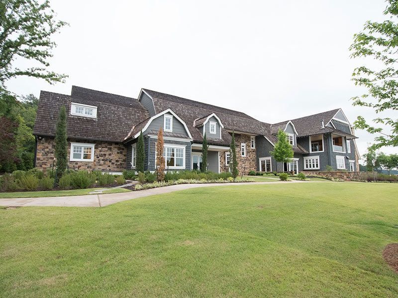 Large gray house with brown roof, green lawn, and trees against a cloudy sky.