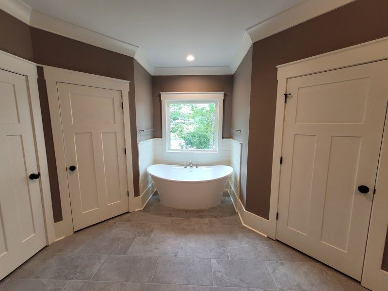 Bathroom with a white bathtub, two white doors, a window, and brown walls. Gray flooring.
