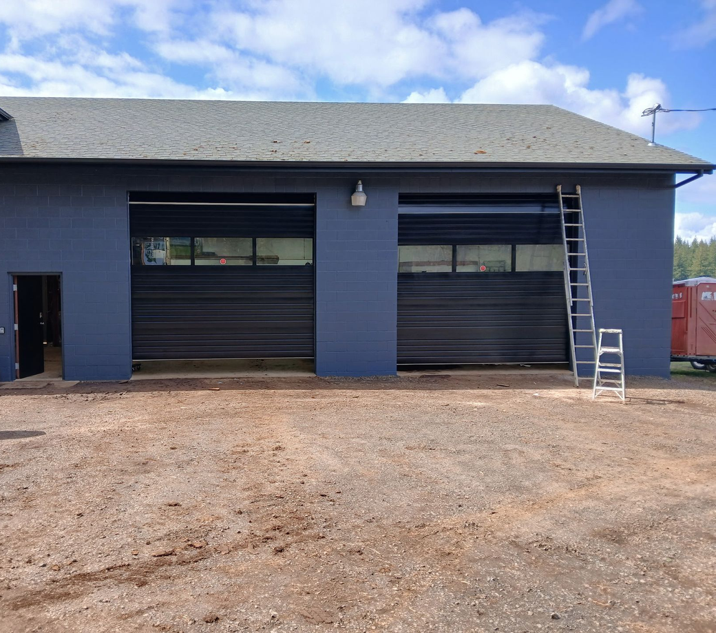 Blue garage building with two roll-up doors, ladder on the right, and gravel foreground