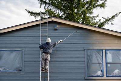 Person on a ladder painting the gray exterior of a house with a roller