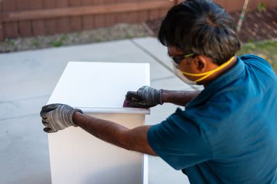 Person wearing gloves and a mask sanding a white board outdoors on a patio.