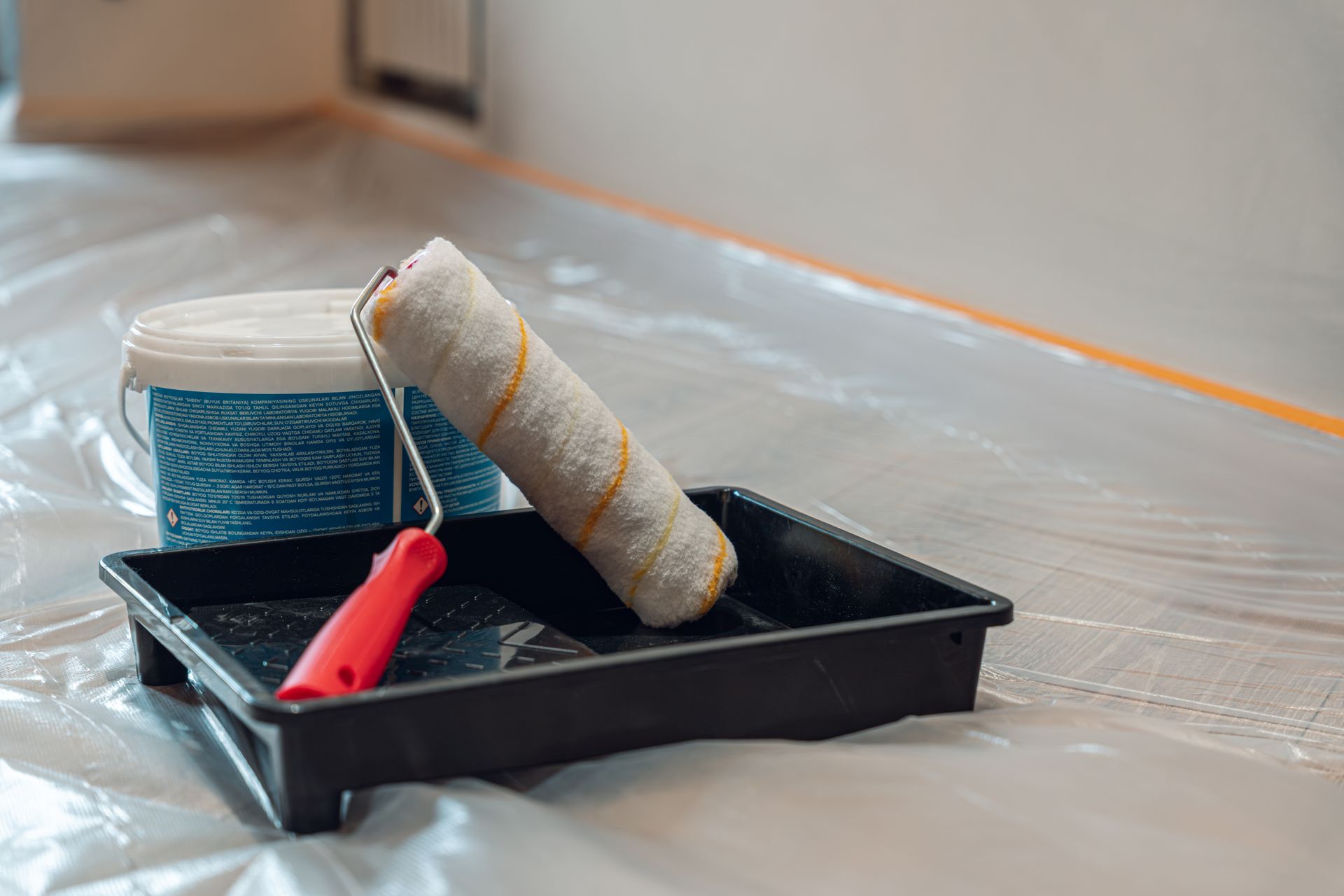 Paint roller tray with blue paint, roller, and red handle on a plastic-covered floor.