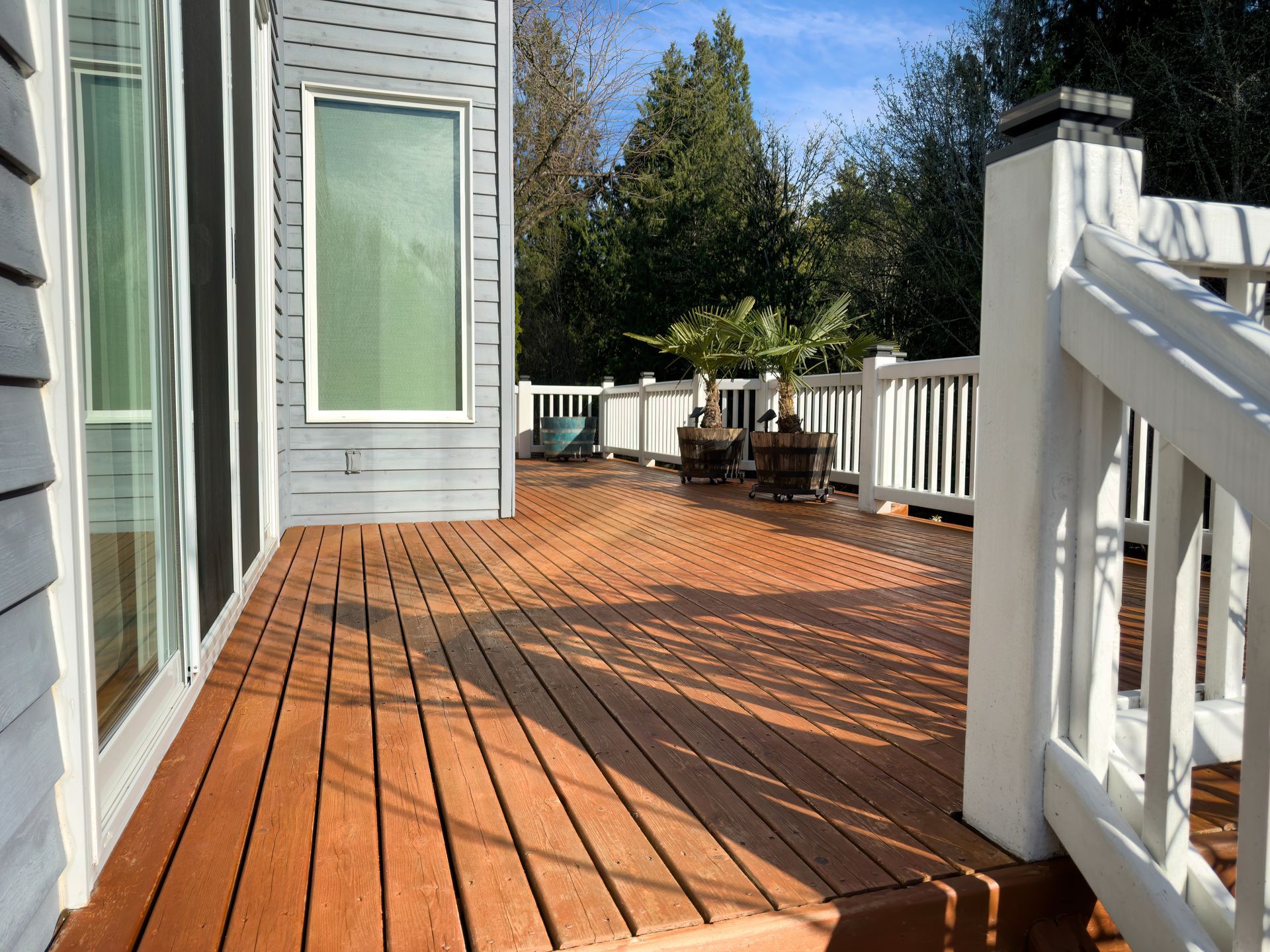 Wooden deck with white railings beside a gray house, lined with trees and patio chairs in the distance