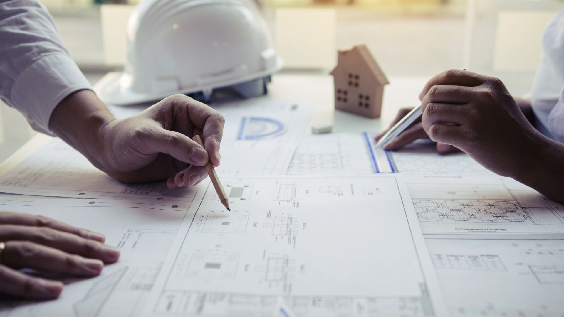 Two people's hands pointing at blueprints on a table with a model house and a hard hat nearby.