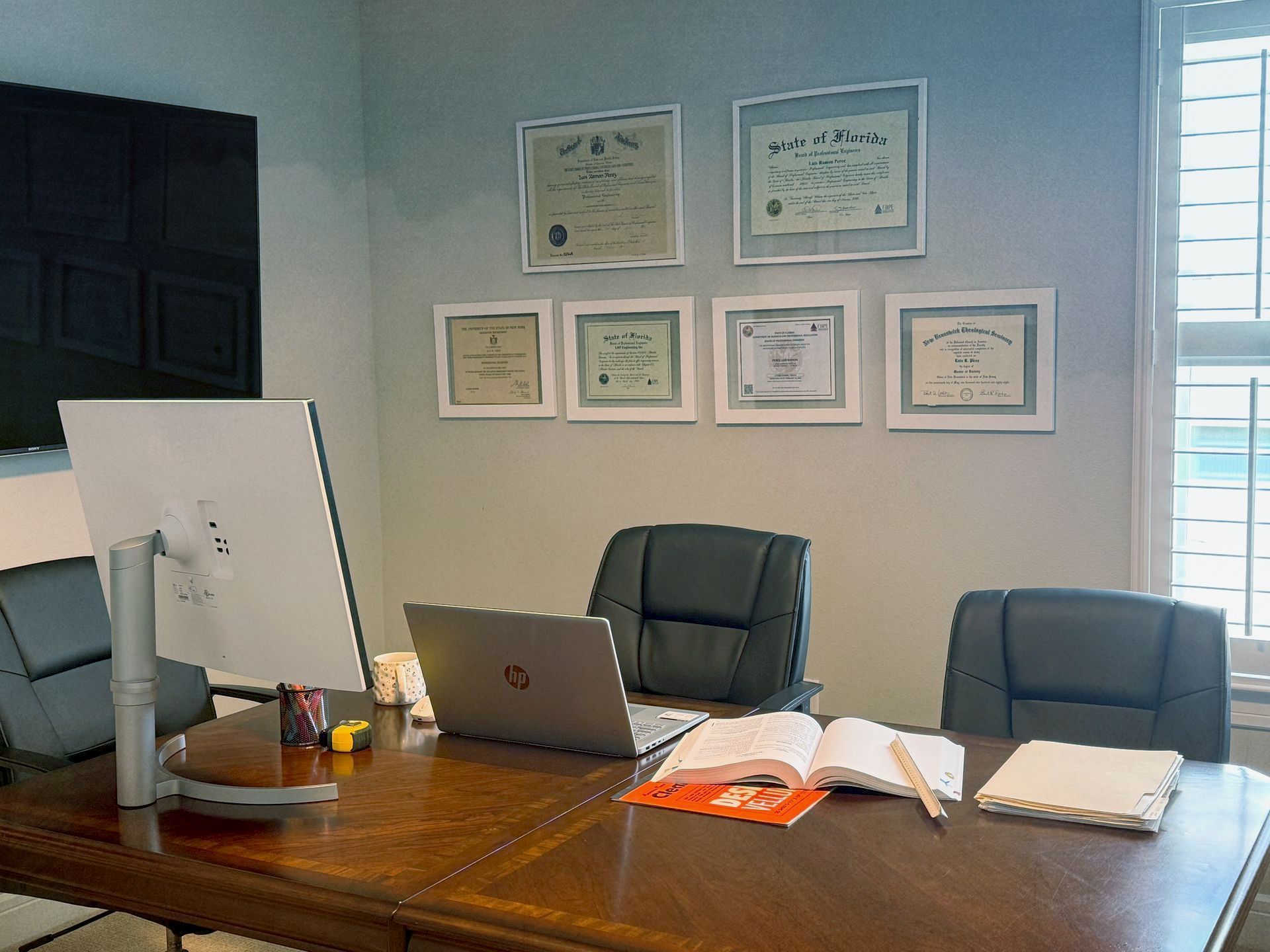Office desk with laptop, monitor, leather chairs, and framed certificates on wall.