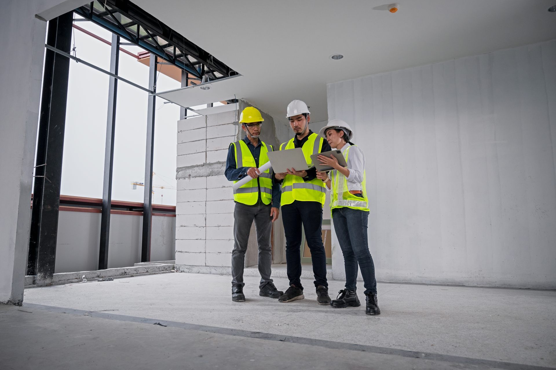 Construction workers in hard hats and vests review plans on a laptop in a building interior.