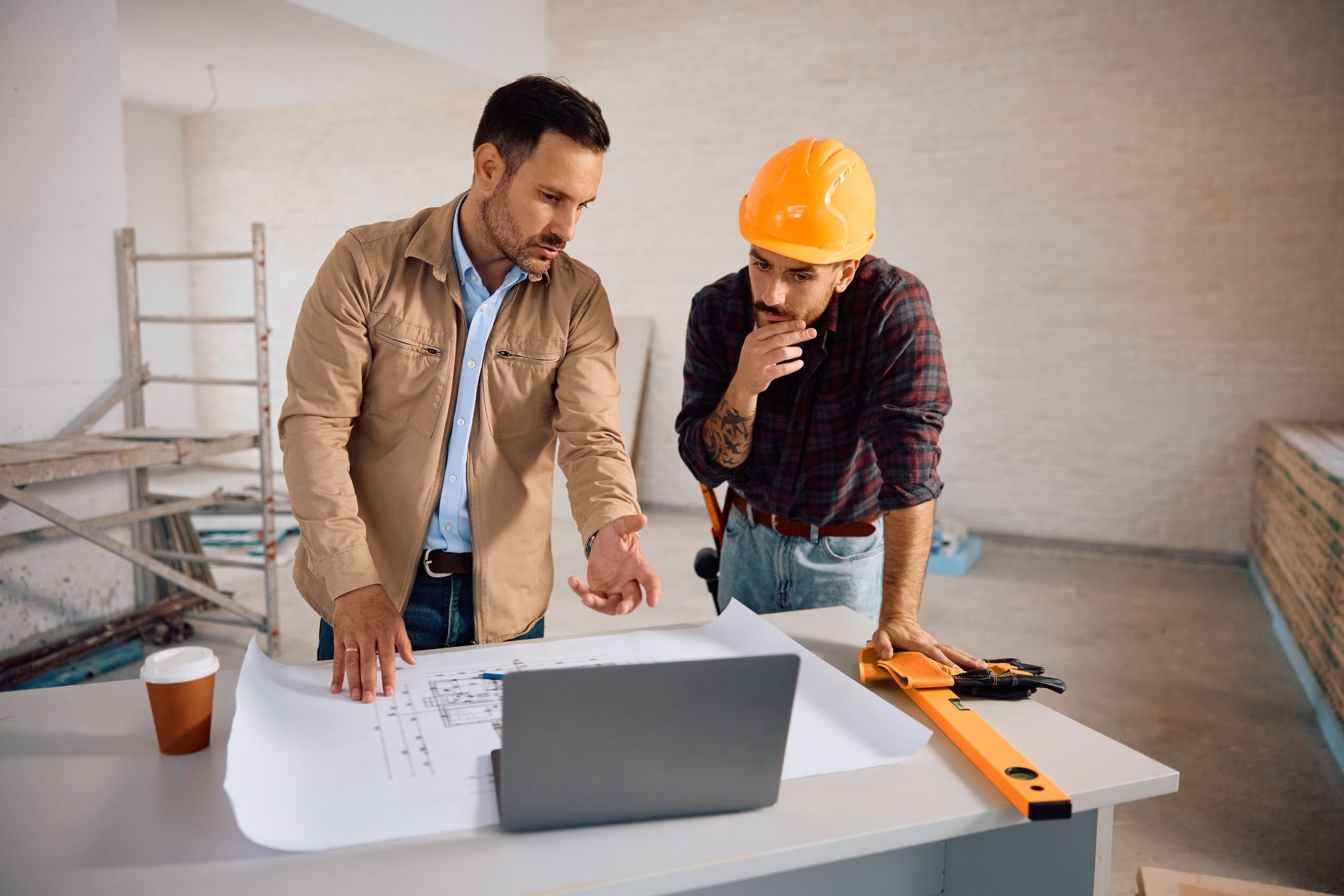 Two men looking at blueprints and laptop at a construction site. One man in a hard hat, the other in a tan jacket.