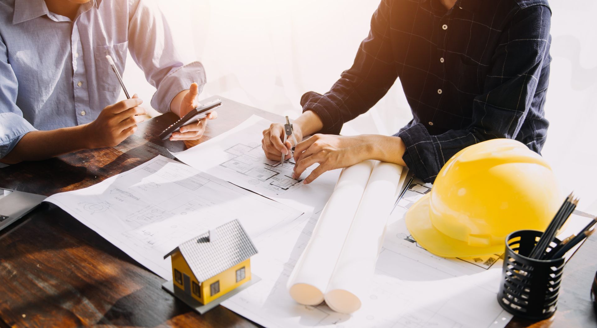 Two people reviewing blueprints at a table with a model house and a yellow hard hat.
