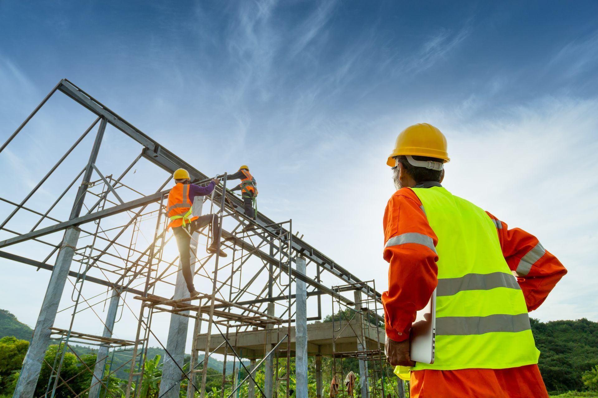 Construction workers building a steel frame structure under a blue sky, one wearing a yellow safety vest.