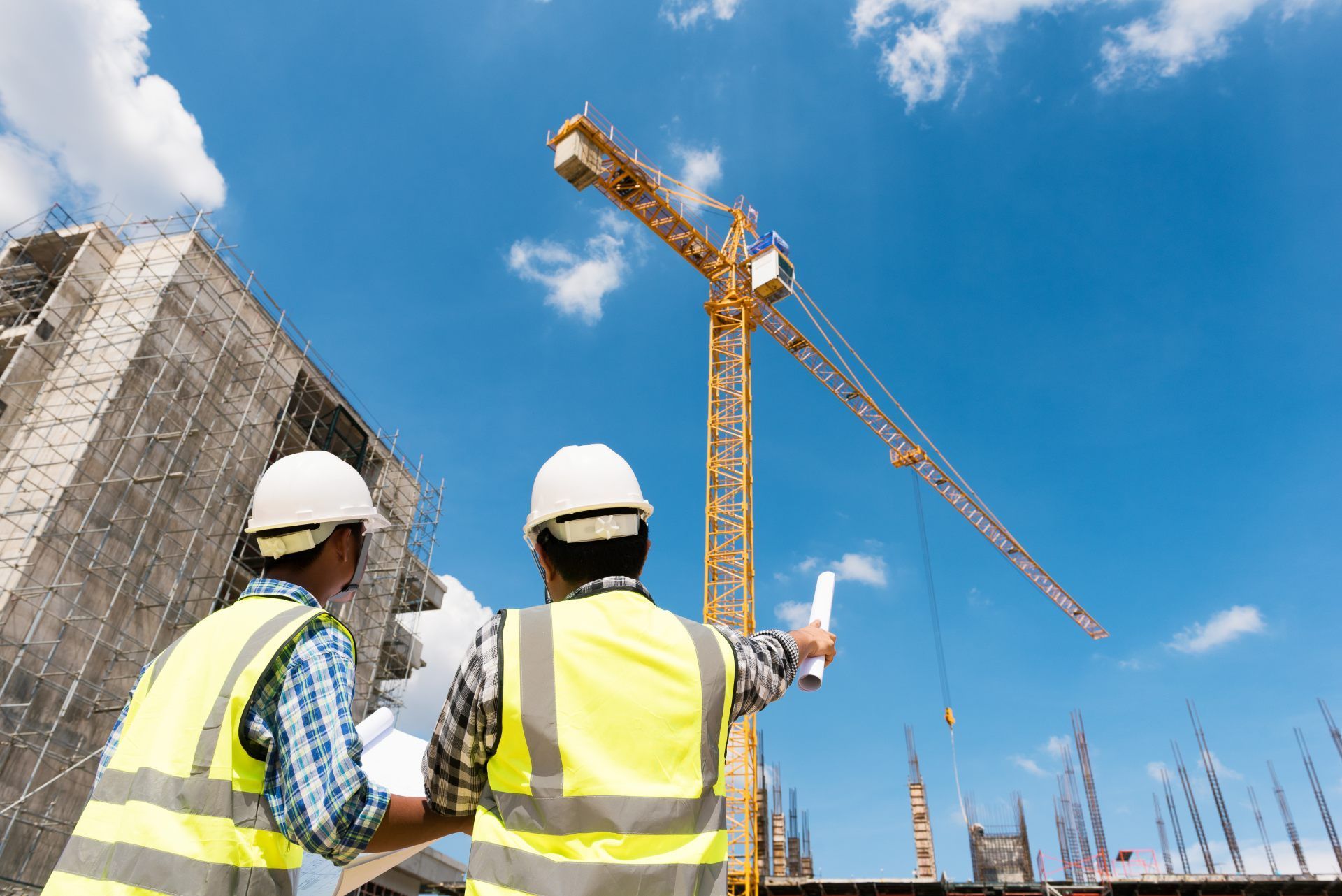 Two construction workers in safety vests and helmets review blueprints at a building site with a crane.
