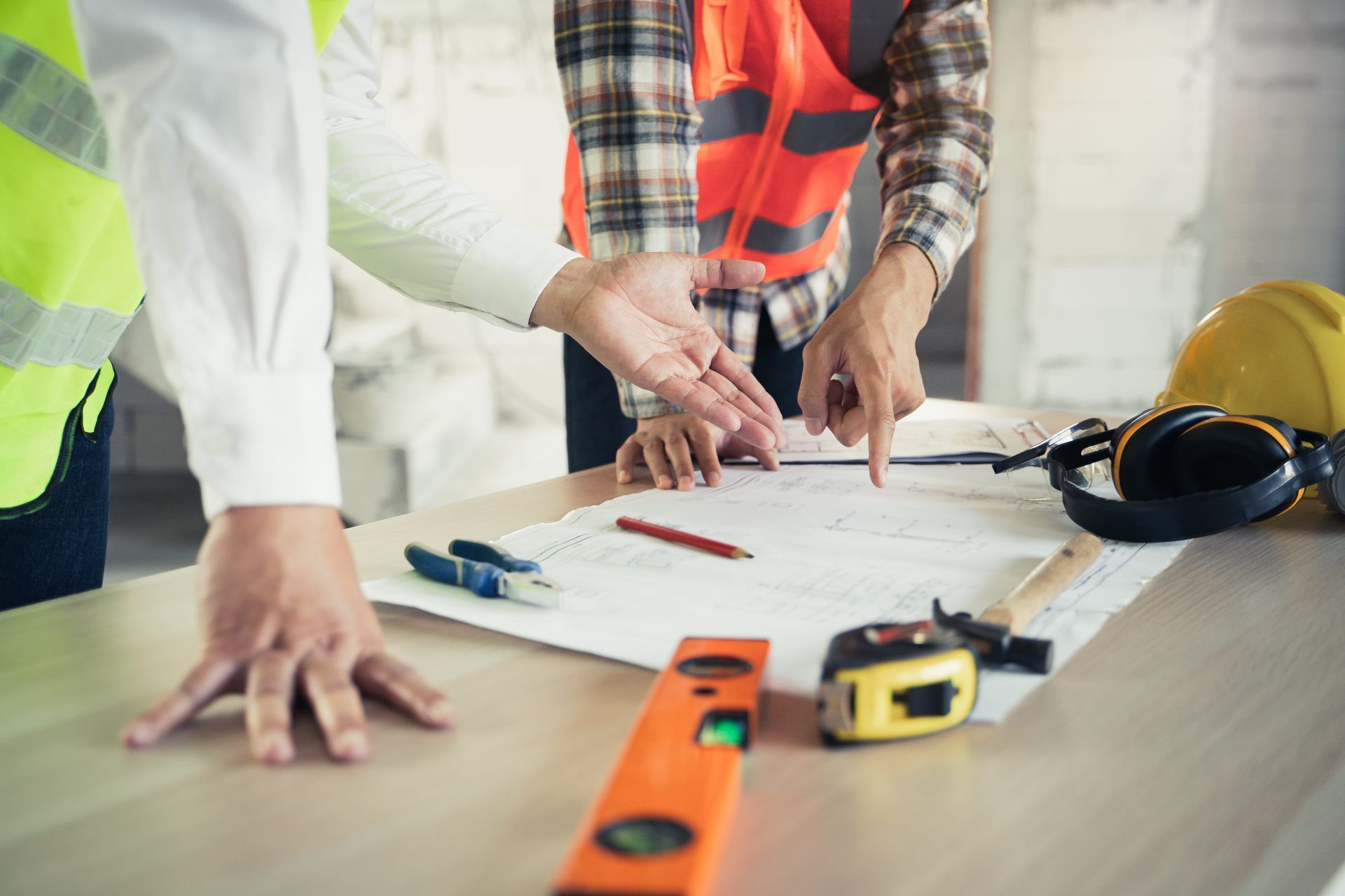 Construction workers reviewing blueprints, pointing at details on a table with tools like a level and hammer.