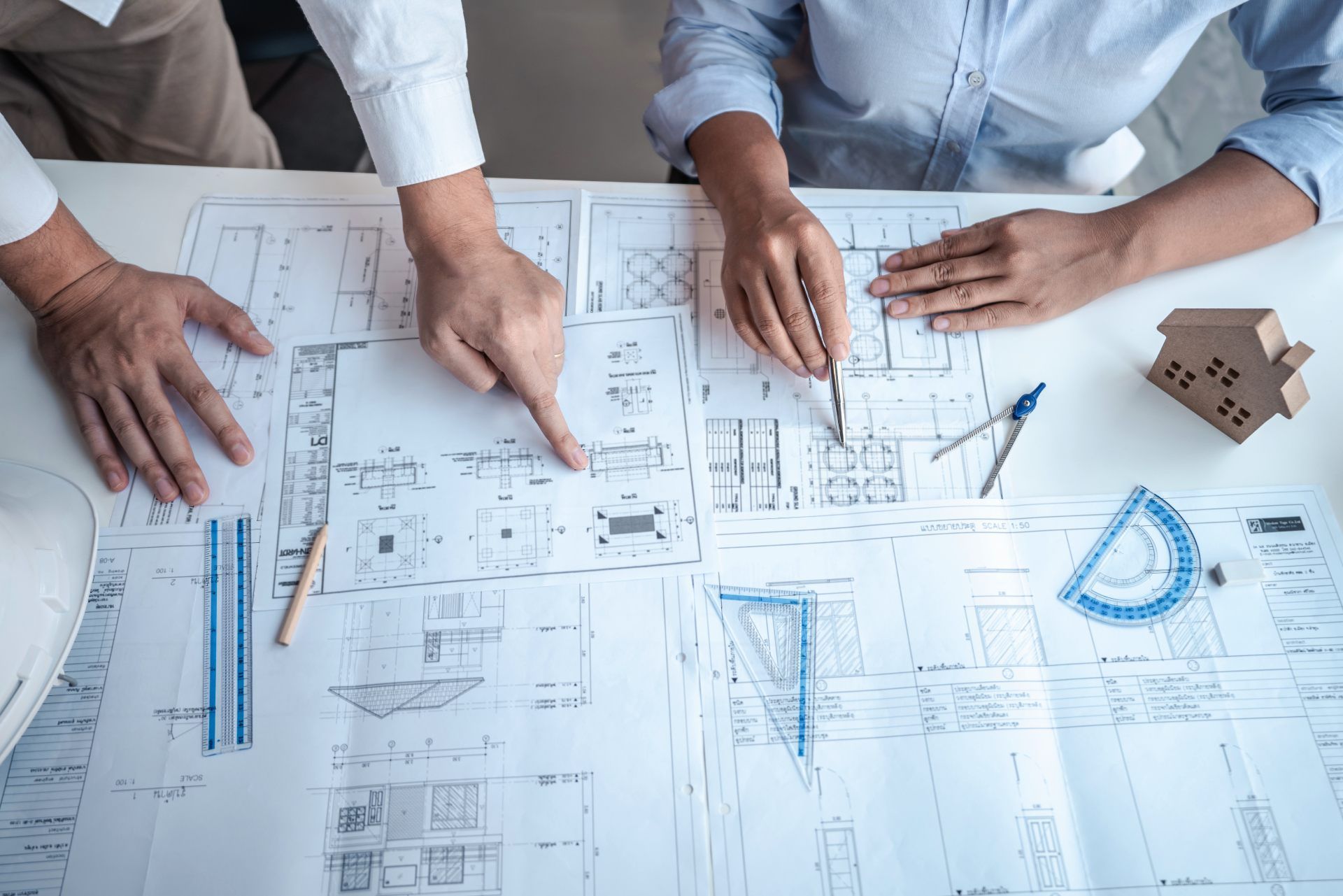 Two people reviewing architectural blueprints at a desk, pointing and using drafting tools.