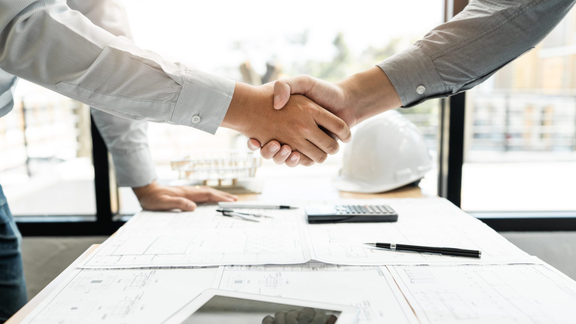 Two people shaking hands over a table with blueprints, a calculator, a pen, and a hard hat.