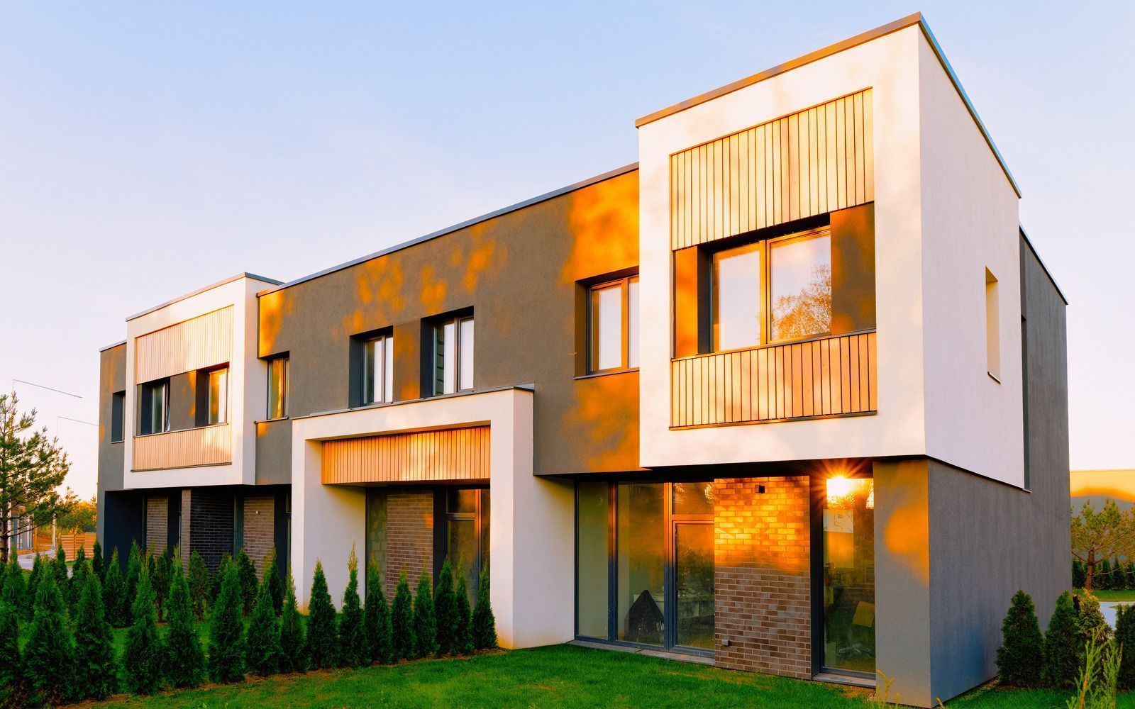 Modern two-story townhomes with white and gray exteriors, wooden accents, and glass windows. Green lawn in front.
