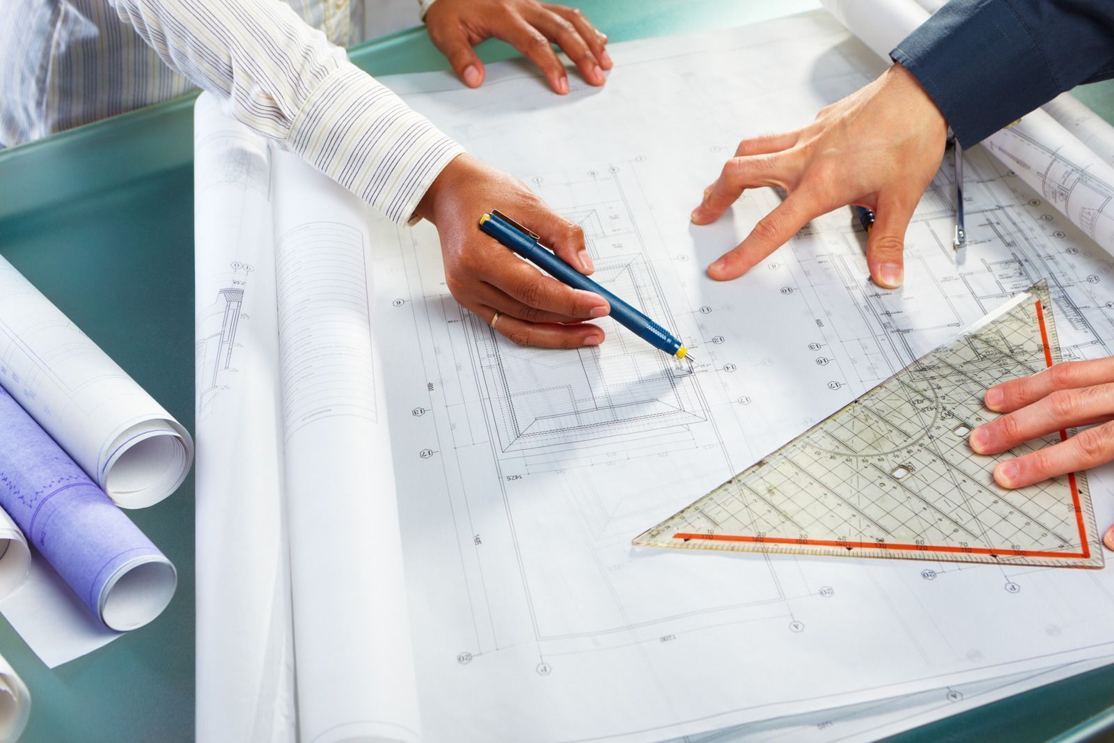 Hands pointing at architectural blueprints, a triangle ruler, and rolled plans on a table.