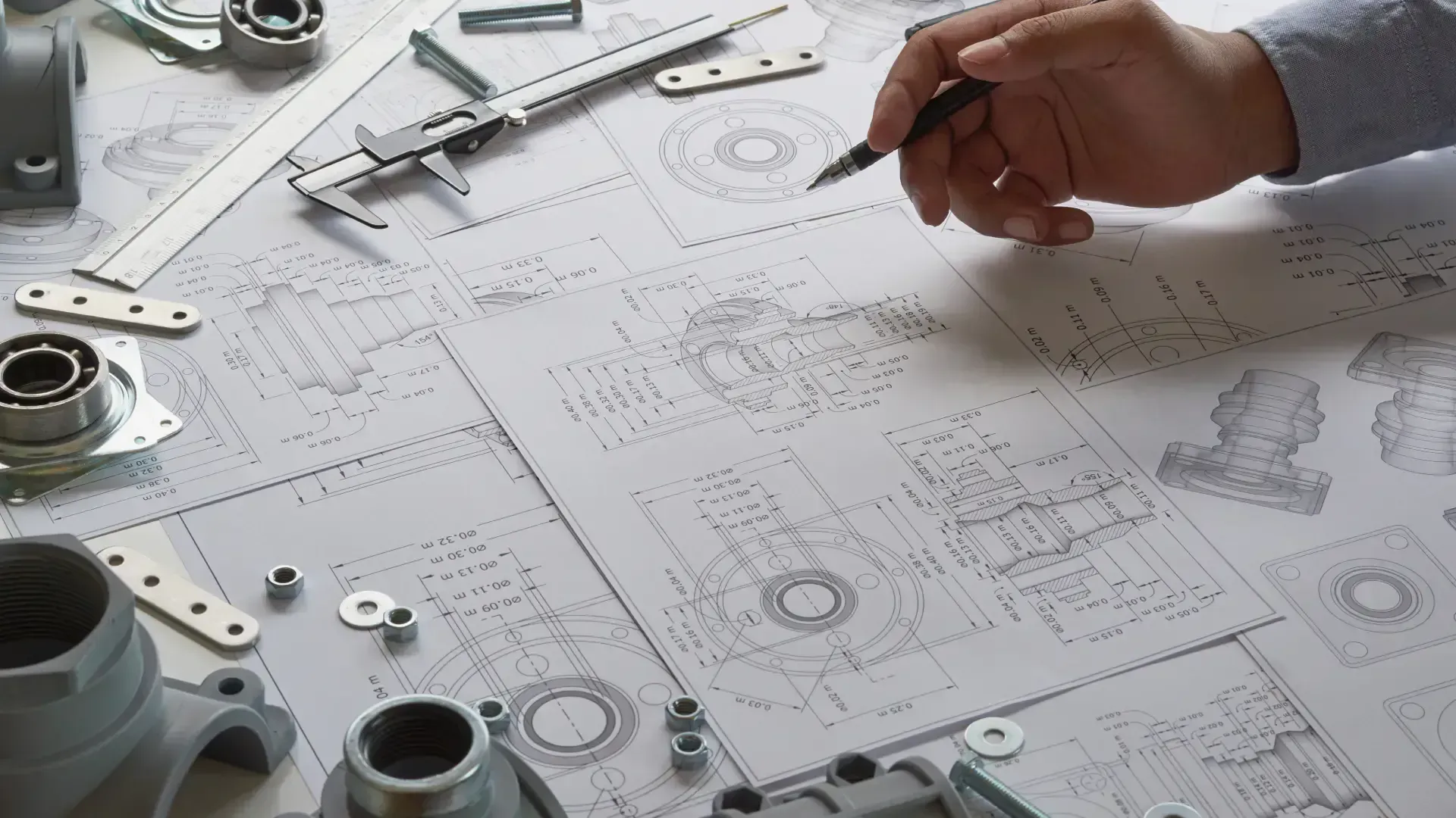 Hand sketching on blueprints with mechanical parts nearby.