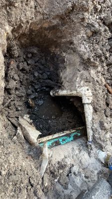 Excavated dirt pit reveals broken PVC pipes and fittings, possibly a plumbing repair site.