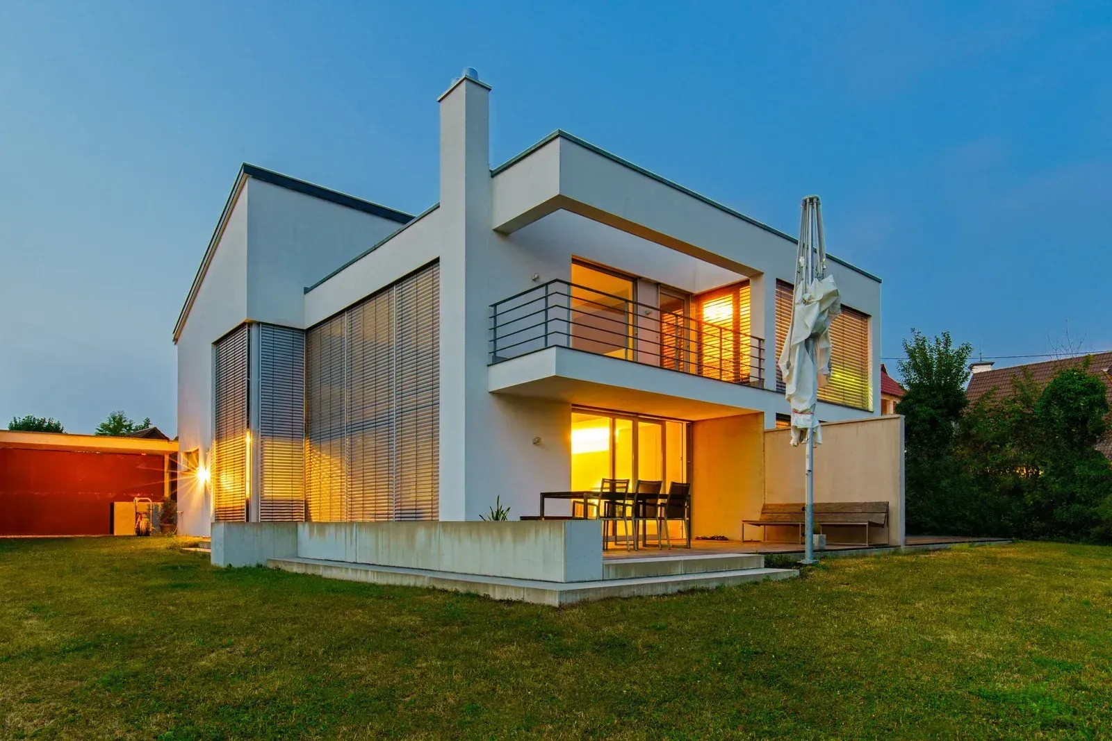Modern white two-story house with balcony and outdoor seating, illuminated by warm interior lights, dusk.