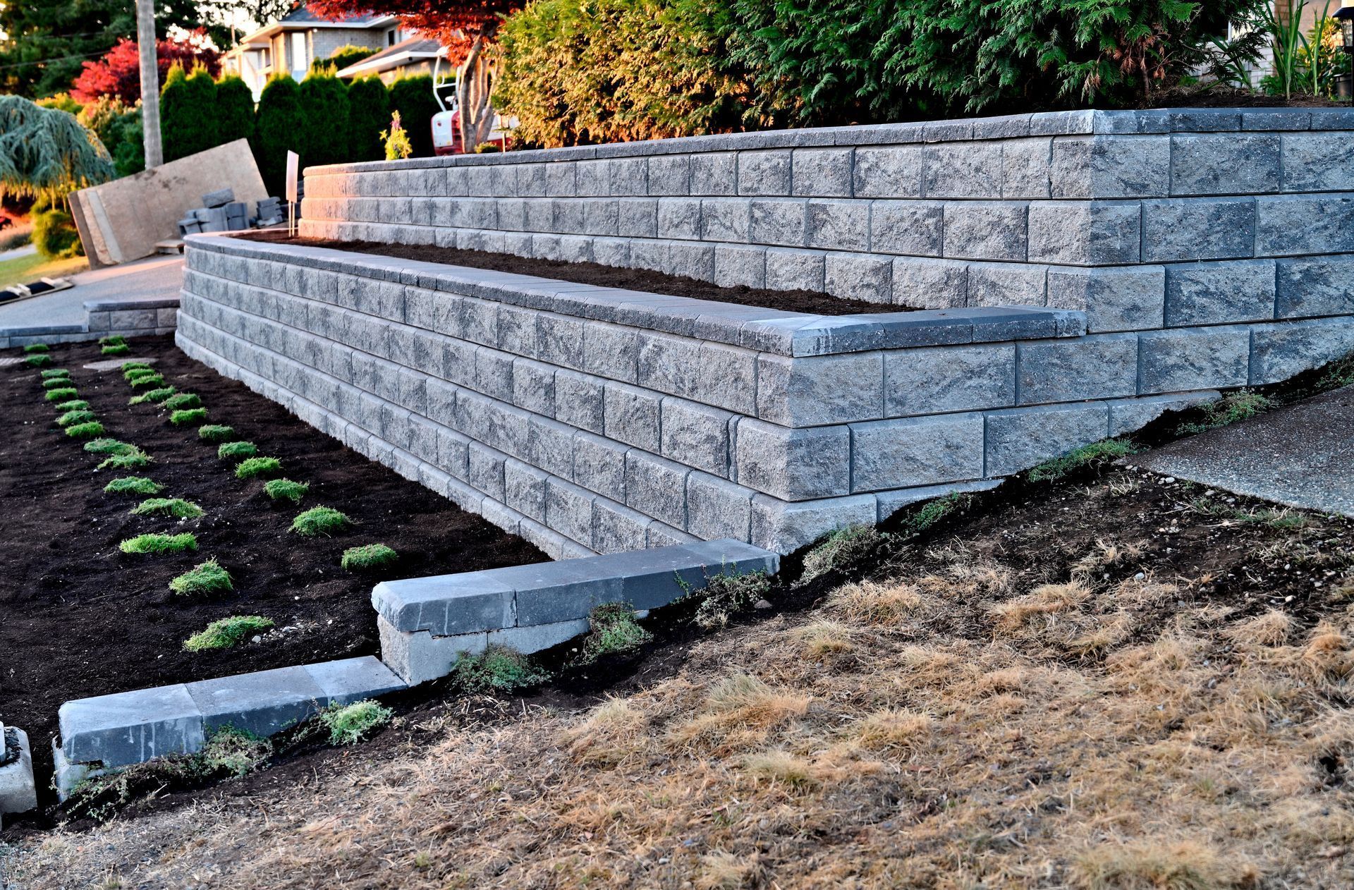 Gray brick retaining wall with tiered garden beds. New plants in beds, slope in foreground.